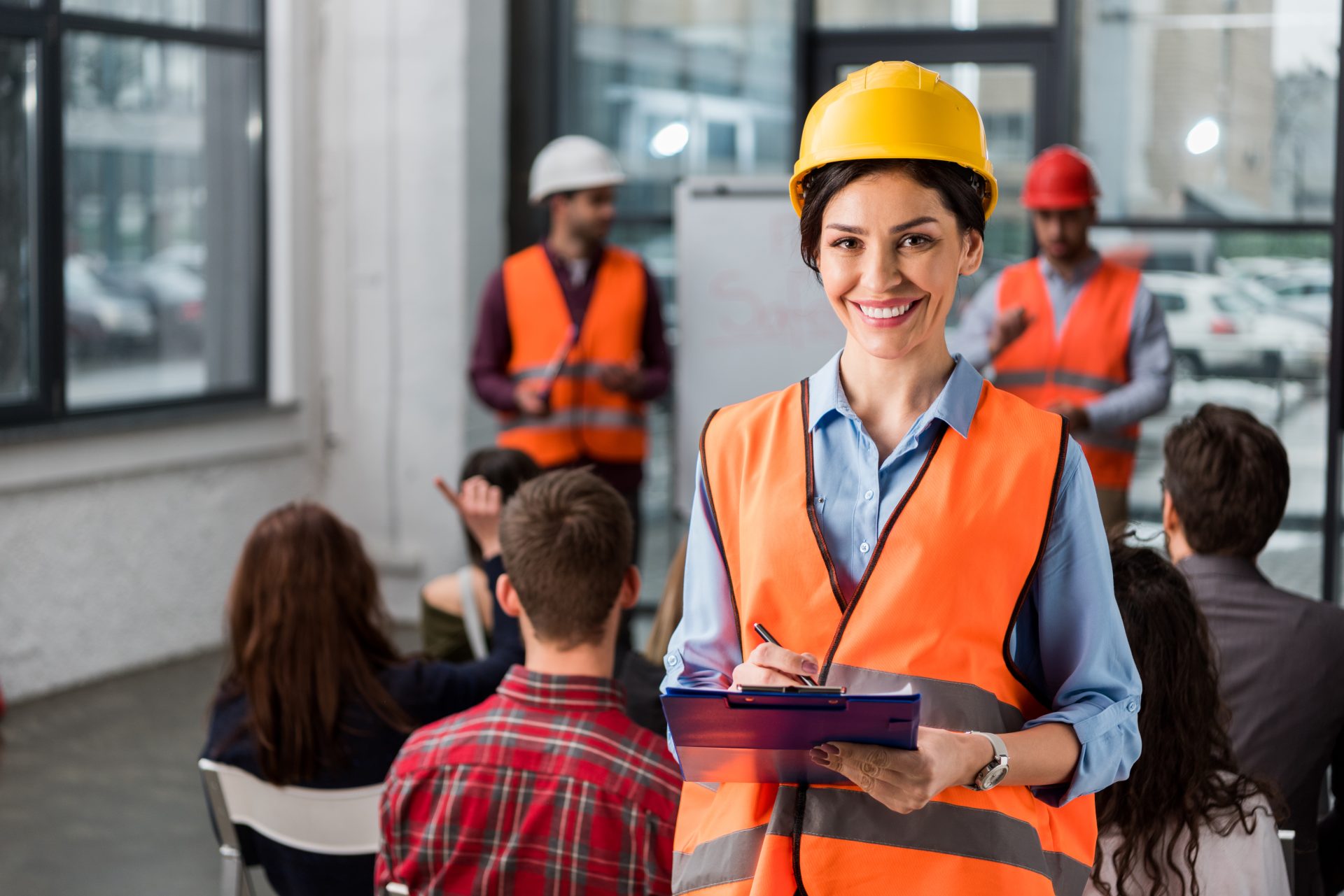 A woman in a yellow hard hat and orange safety vest smiles with a clipboard in hand. Behind her, colleagues in similar attire conduct a safety training session, illustrating the importance of OSHA reporting in maintaining workplace safety.