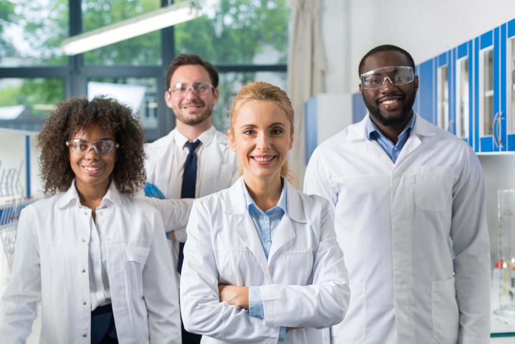 Smiling Group Of Scientists In Modern Laboratory With Female Leader, Mix Race Team Of Scientific Researchers In Lab Wearing White Coats And Protective Glasses
