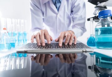A scientist in a laboratory uses a laptop for work, surrounded by lab equipment, test tubes, and safety gear on the table.